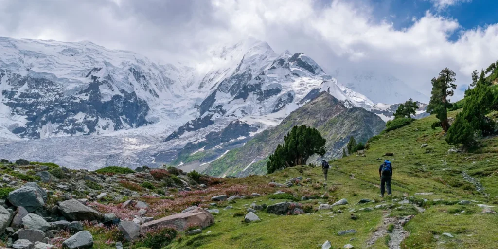 Two trekkers hiking through green alpine meadows with snow-capped Himalayan peaks in the background — best time for an Uttarakhand trek is September to October.