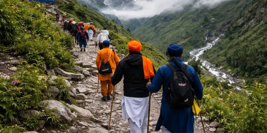 Pilgrims, many Sikh devotees wearing colorful turbans and carrying walking sticks, trek along a rocky mountain path toward Hemkund Sahib, surrounded by lush green valleys and mist-covered snow-capped Himalayan peaks.